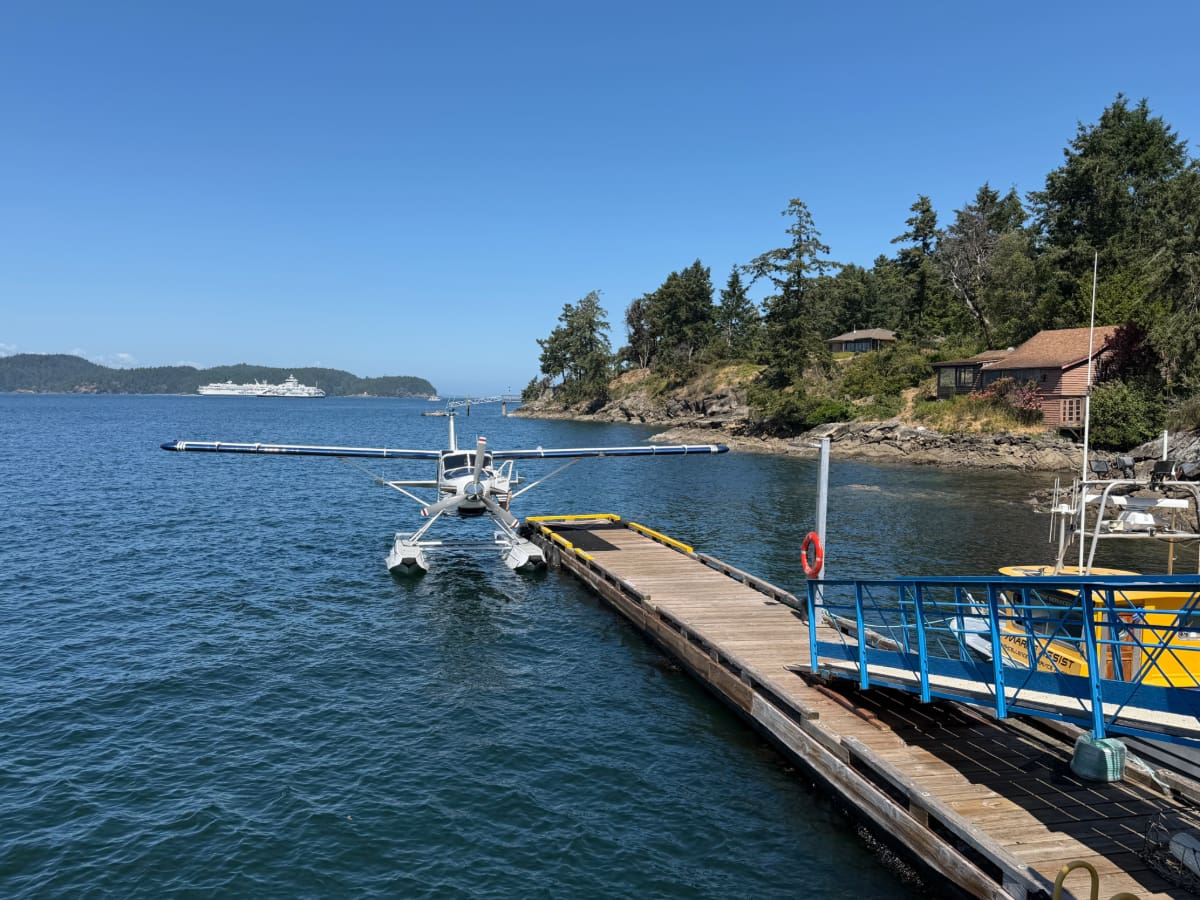 A seaplane docked at a wooden pier on a calm bay, with a yellow boat, rocky shoreline, trees, and houses in the background under a clear blue sky.